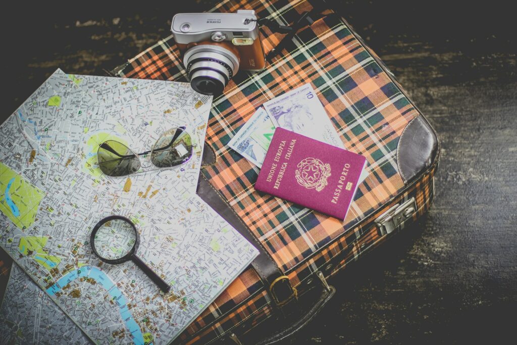 Imagem de uma mala xadrez em preto e laranja, sob ela está uma câmera fotográfica, um passaporte, algumas notas de dinheiro, um óculos de sol, uma lupa e um mapa