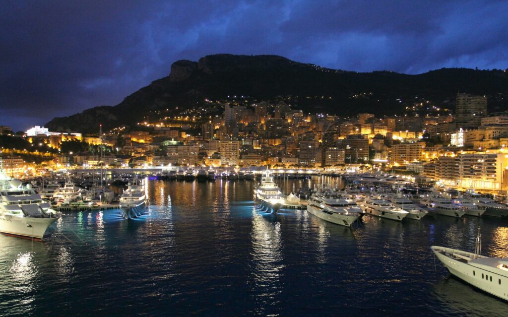 Região de Monte Carlo, num porto de noite com barcos parados e as luzes acesas, ao fundo, a cidade iluminada, para representar o seguro viagem para Mônaco