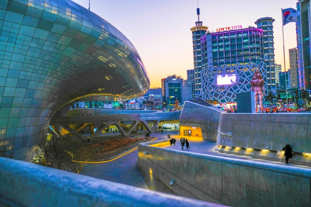 O complexo arquitetônico Dongdaemun Design Plaza na Coreia do Sul, um prédio com curvas ondulares inteiro de metal, refletindo as luzes que estão ao redor com uma ampla rampa na sua entrada com pessoas caminhando, para representar o seguro viagem para Coreia do Sul