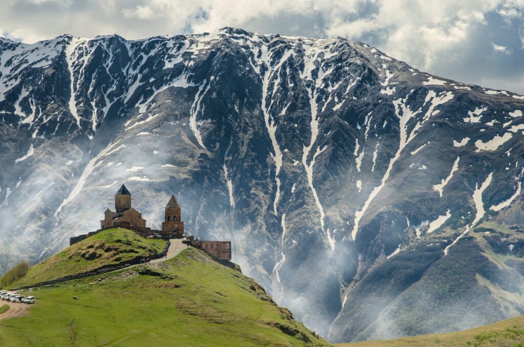 Igreja da Trindade Gergeti em Kazbegi na Geórgia, uma construção na beira de uma montanha que, ao fundo, é quase coberta por uma montanha infinitamente maior e cheia de neve, para representar o seguro viagem para Geórgia
