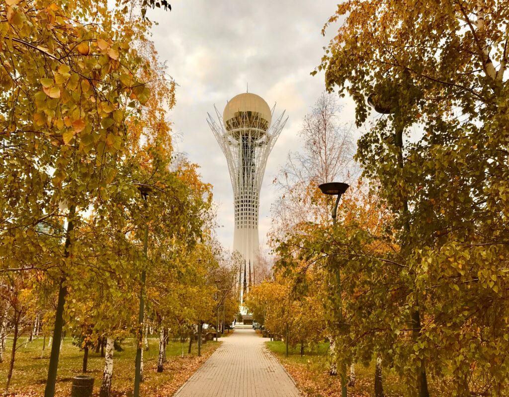 vVisão de um parque com árvores em tons de amarelo que formam um caminho em direção do Monumento de Baytereks na capital do Cazaquistão, uma torre alta com um globo ao topo, para representar o seguro viagem para o Cazaquistão
