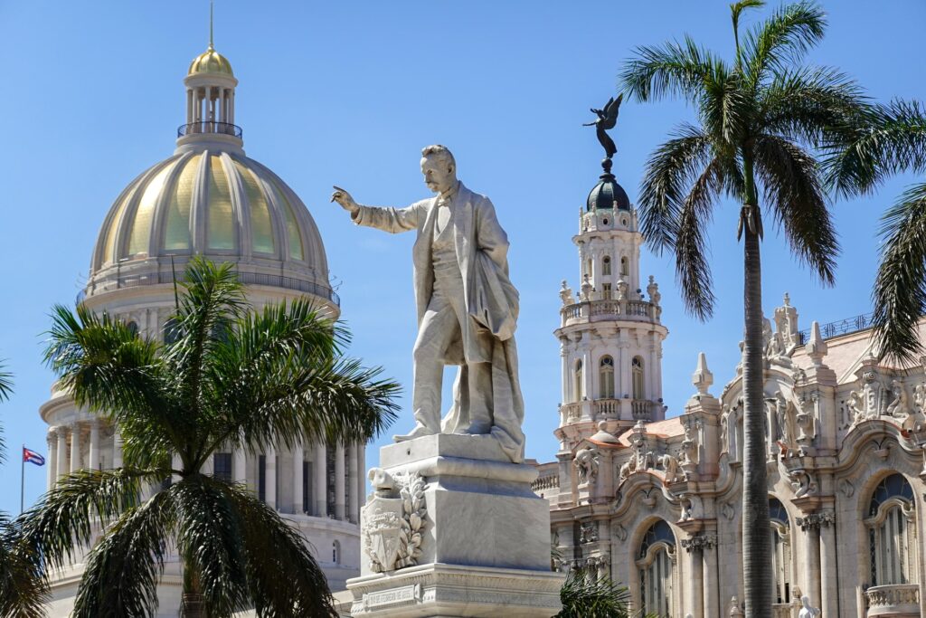 Uma estátua do José Martí, um homem com terno, e um braço erguido com o dedo indicador apontado, ao redor, outros prédios históricos e algumas palmeiras, para representar o seguro viagem para Havana