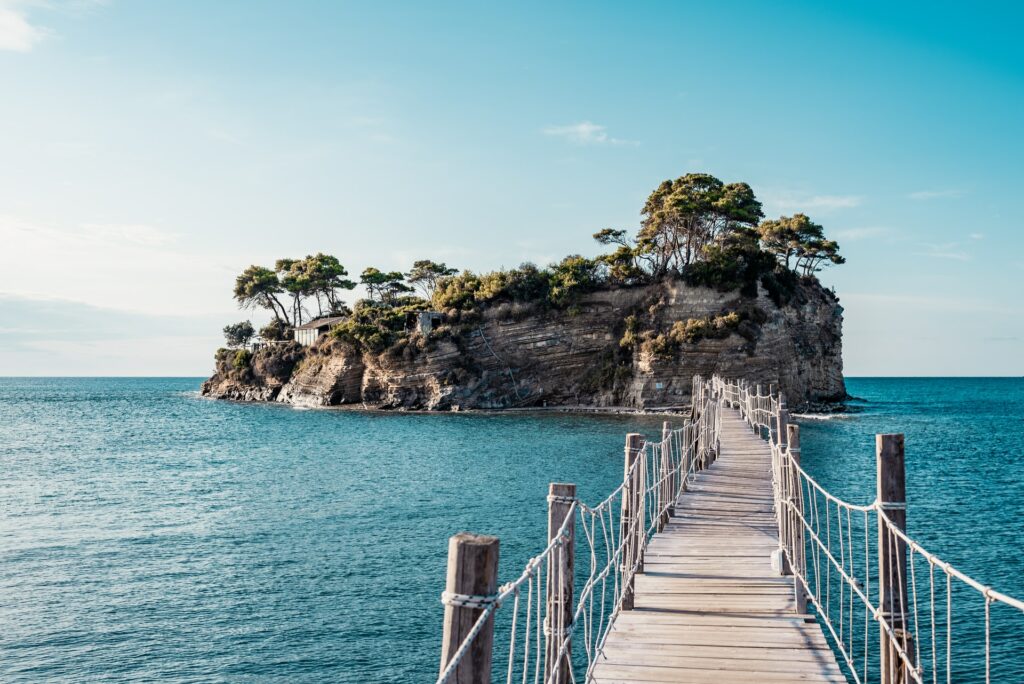 Lagana Beach em Zakynthos com uma ponte que atravessa o mar e leva até uma pequena ilha formada em uma rocha, com algumas árvores, e cercada pelo mar azul claro