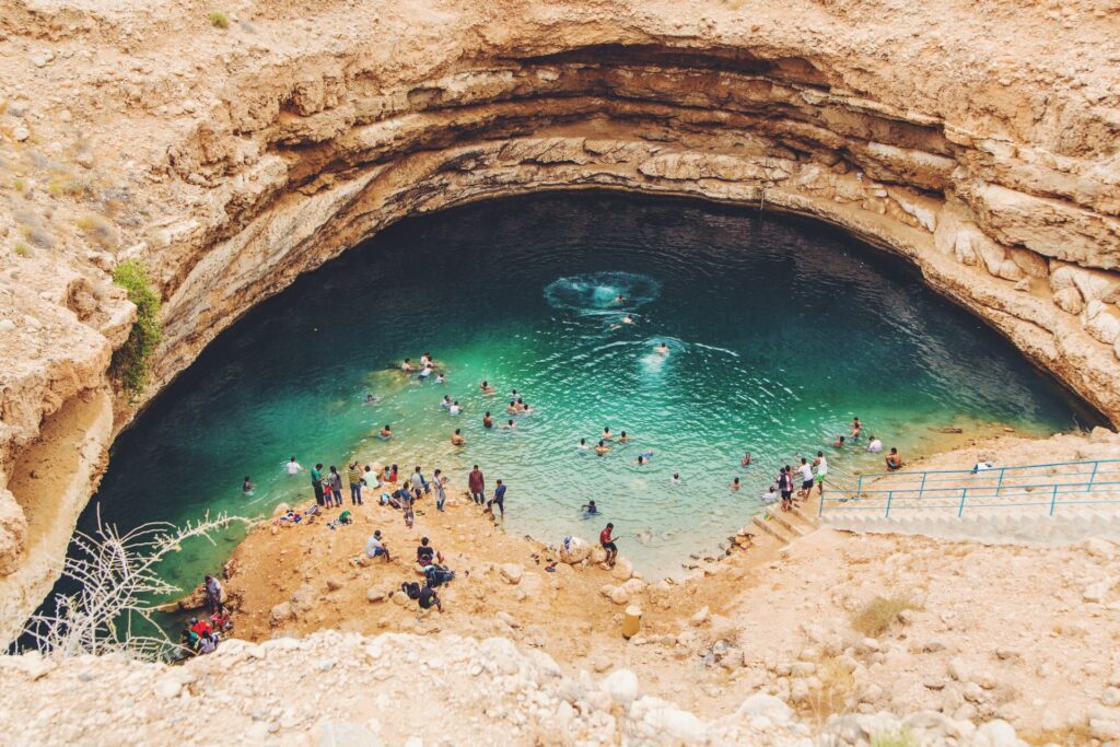 Bimmah Sinkhole em Mascate, capital de Omã, uma lago que se formou no meio de rochas, formando uma espécie de piscina natural, uma escadaria leva até o local de águas verdes, muitas pessoas estão na água e outras sentadas na pedras, para representar o seguro viagem para Omã