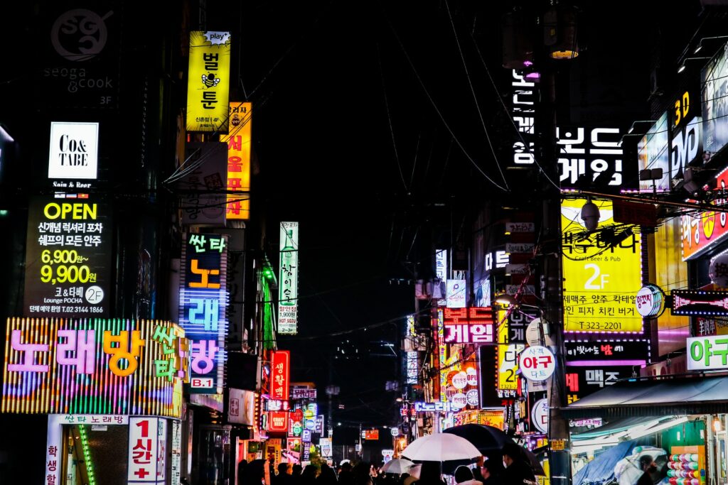 Uma rua de noite totalmente iluminada por placas dos comércios em cores vibrantes, pessoas andando, em Seul na capital da Coreia do Sul