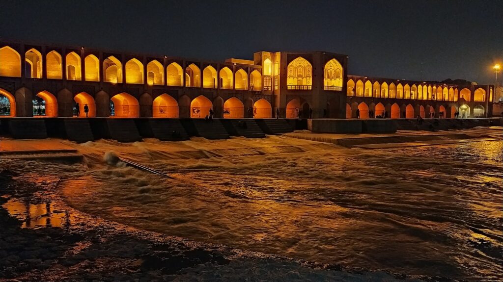 Ponte Khaju em Isafhan no Irã, uma extensa ponte que corta um rio, formato por diversos arcos e completamente iluminada em tons de amarelo e laranja durante à noite, para representar o seguro viagem para o Irã