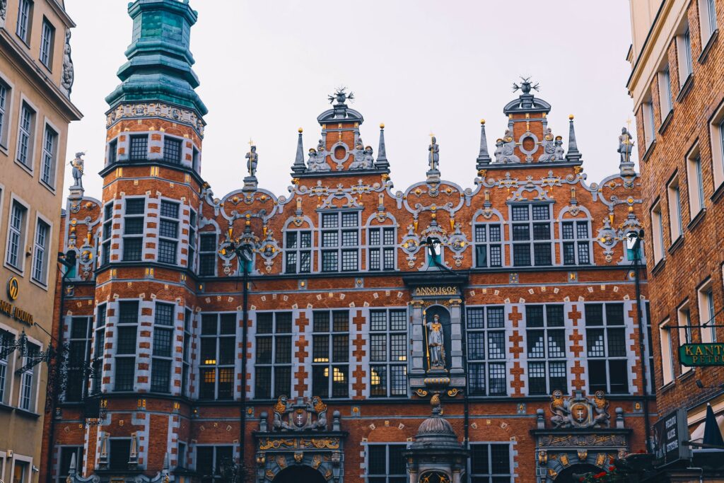 The Great Armoury em Gdansk na Polônia, uma construção antiga em tons laranja, vermelho, azul, cinza, com alguns monumentos de pessoas ao topo das pequenas torres, para representar o seguro viagem para Polônia