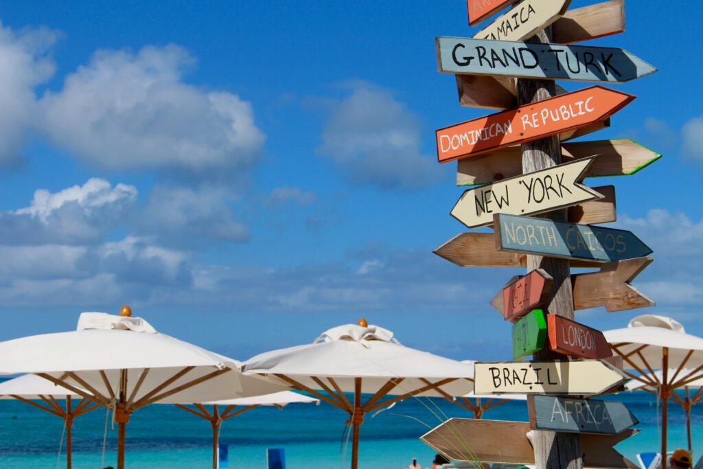 Praia de Grace Bay, muitos guarda-sóis brancos, um poste com placas coloridas com nomes de diferentes países: República Dominicana, Brasil, África, com o azul do mar ao fundo