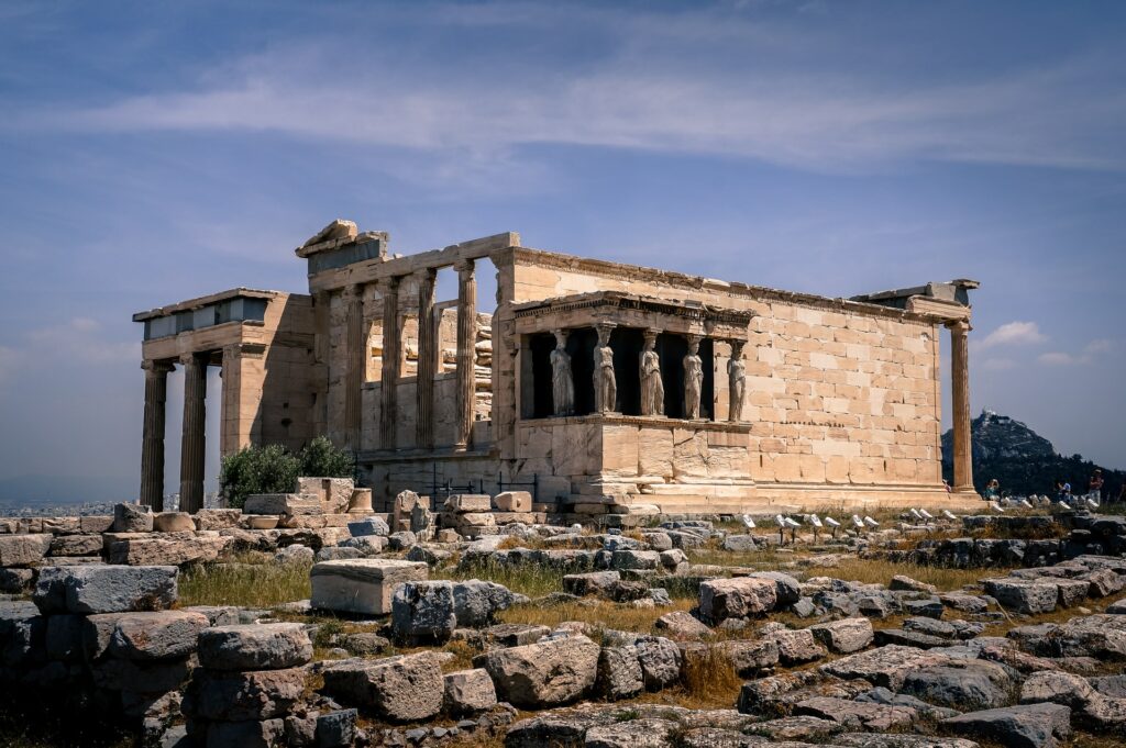 Antigo Templo de Atenas, construído em torno de 525-500 a.C. em Atenas, muitas ruínas ao redor da construção central, colunas altas, na lateral há 5 mulheres esculpidas em pé, sustentando uma parte da construção, para representar o seguro viagem para Atenas