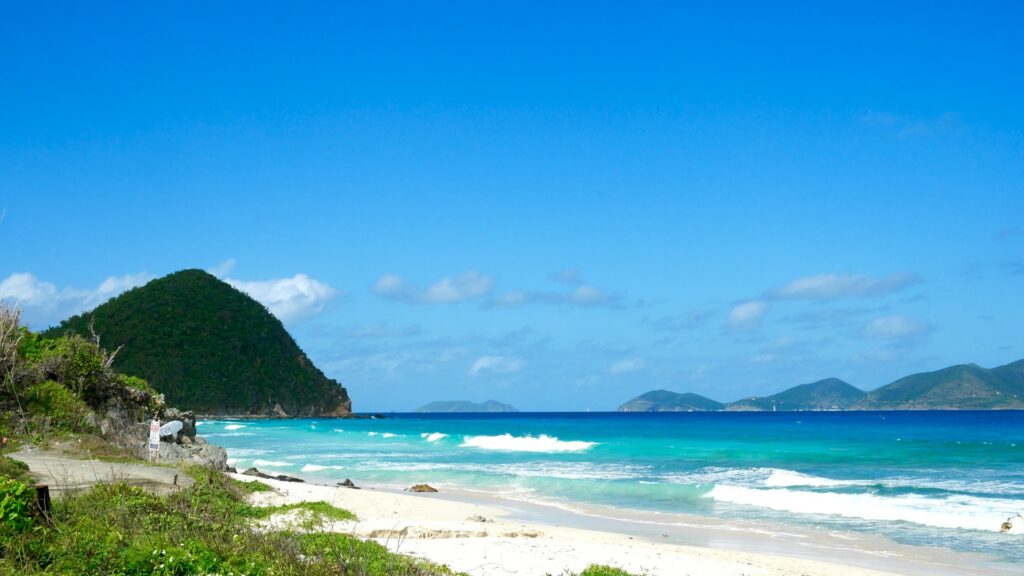 Tortola, uma praia quase deserta com areia branca, mar em tons de azul claro e escuro, ao fundo do mar, é possível ver diversas montanhas localizada nas ilhas virgens britânicas