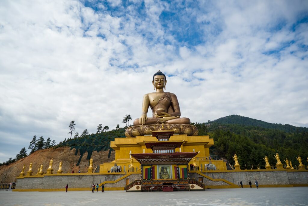 Mosteiro Dzong com uma enorme estátua de Buda segurando um pote sob uma construção em tons de dourado e com um enorme pátio logo a frente com pessoas caminhando, para representar o seguro viagem para Butão