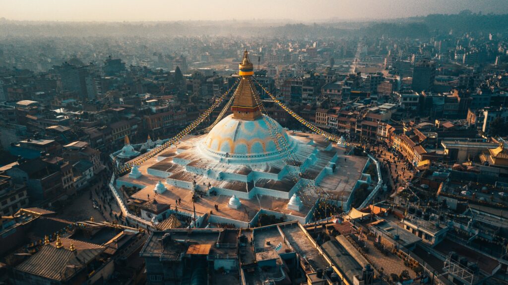Templo budista de Boudhanath Stupa em Catmandu, um enorme templo em tons de azul e branco montado como se fosse uma lona, ao topo um pequeno monumento em tons de dourado, e ao redor da construção está a cidade, para representar o seguro viagem para o Nepal