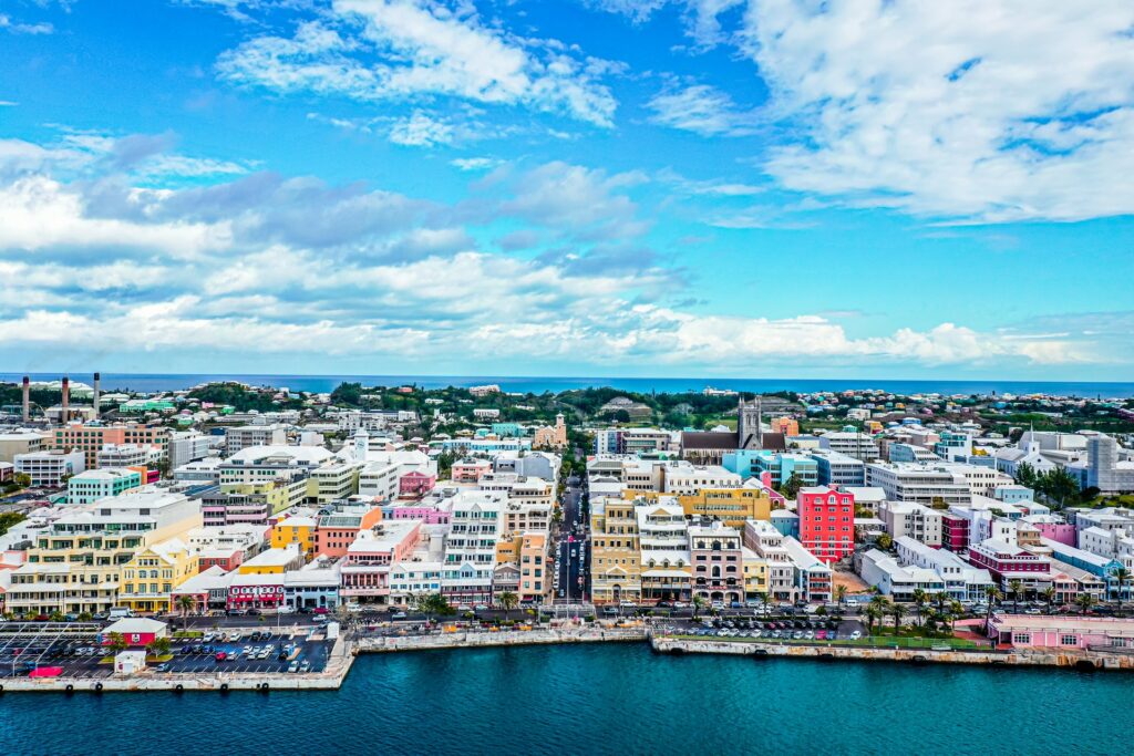 Hamilton, capital de Bermudas, um local com prédios coloridos e antigos, vermelhos, rosas, amarelos, uma avenida passa na beira no mar, ao fundo, é possível ver algumas árvores e, depois, o mar novamente, para representar o seguro viagem para Bermudas