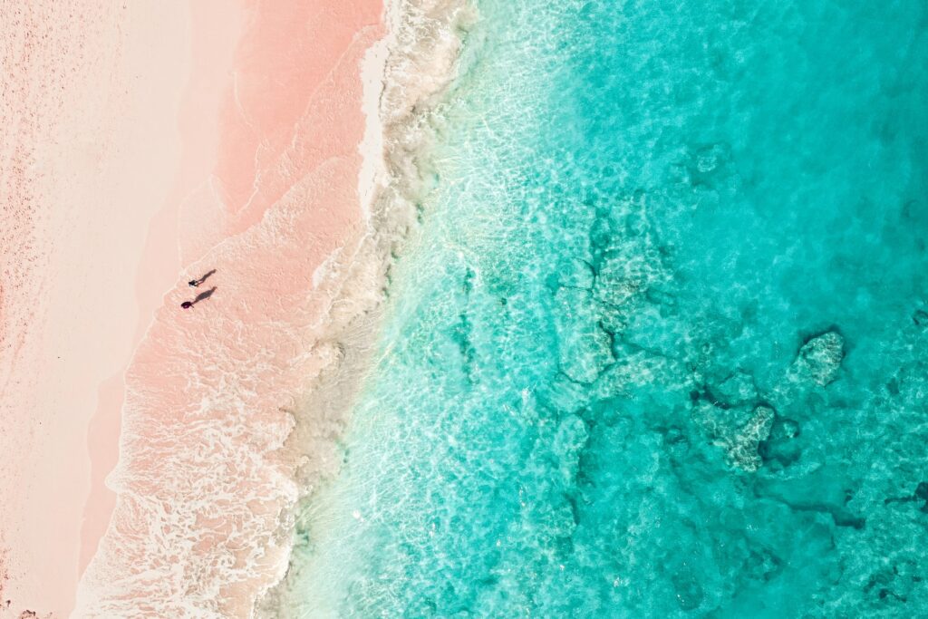 Praia Elbow que tem a areia em um tom de rosa claro que chama atenção, o mar é tão claro que é possível ver o fundo, duas pessoas estão paradas observando o mar, para representar o seguro viagem para Bermudas