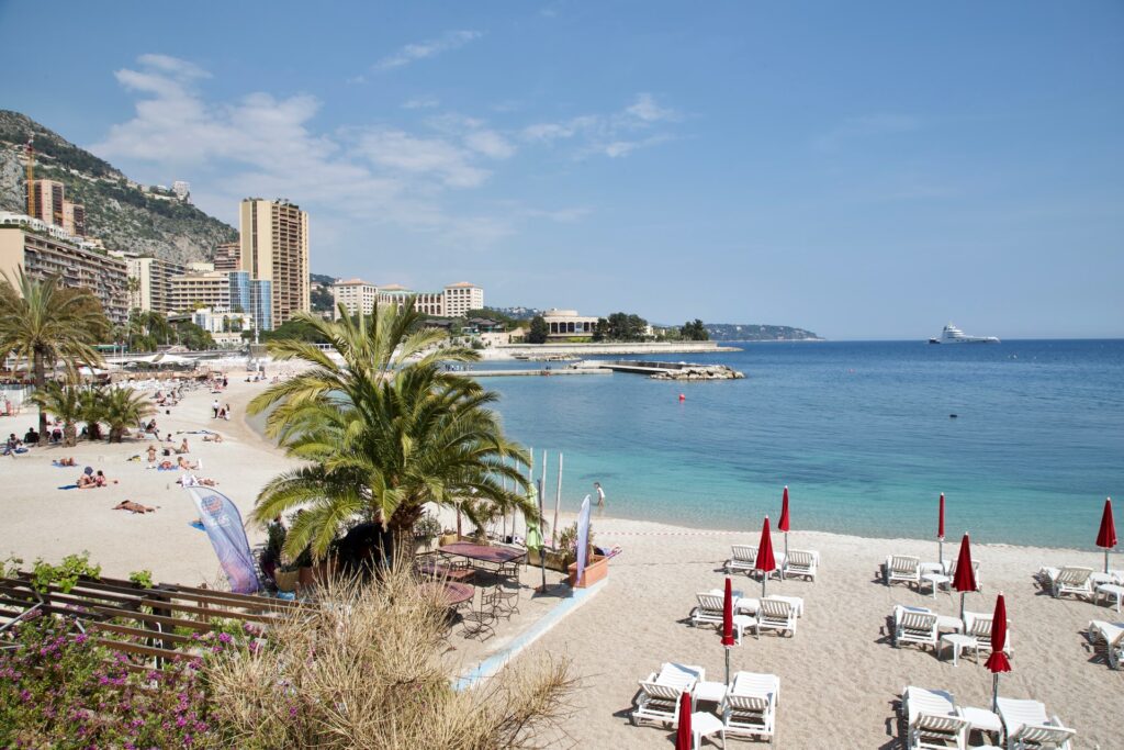 Praia Larvotto em Mônaco, uma praia com água azul bem clara, aréia igualmente clara, algumas pessoas deitadas pegando sol, ao fundo da praia, prédios luxuosos e, mais ao fundo, as montanhas, para representar o seguro viagem para Mônaco