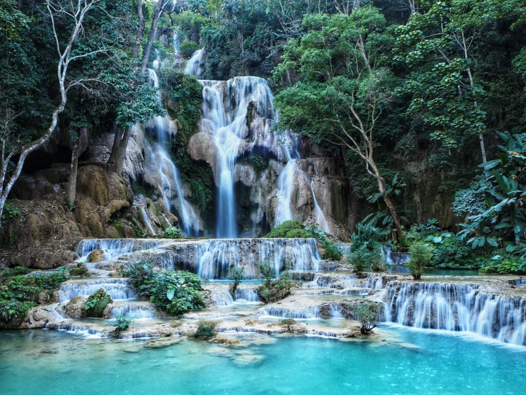 Cachoeiras de Kuang Si em Laos, diversas quedas d'águas em tons de azul claro por entre rochas, cercadas de natureza, que formam um lago ao final da queda, para representar o seguro viagem para Laos
