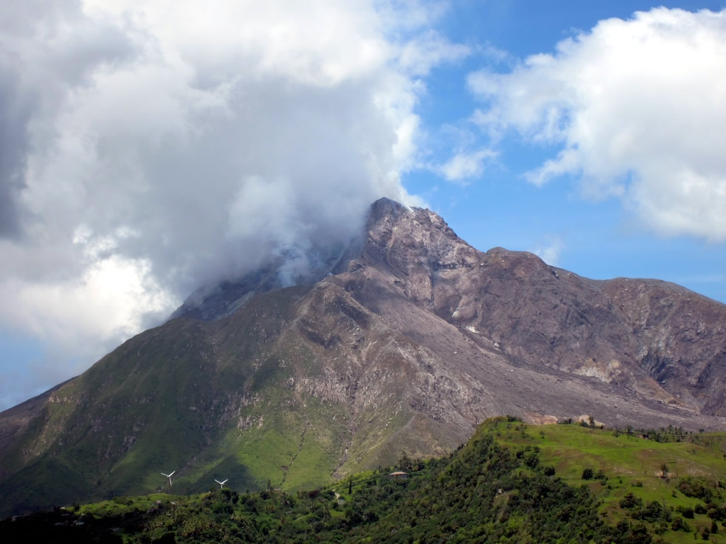 Vulcão Soufrière Hills, uma vulcão composto de enormes rochas, coberto por uma nuvem cinza passageira, ao pé do vulcão, muita vegetação e árvores, para representar o seguro viagem para Montserrat