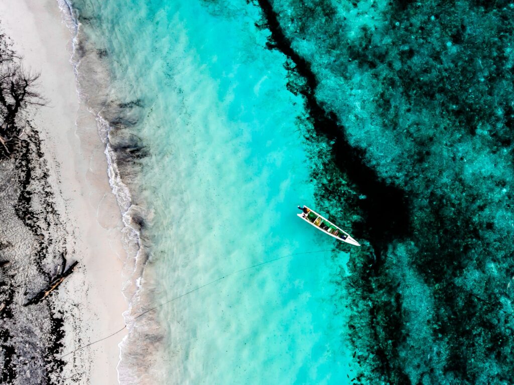 Acrema Beach em Timor Leste, uma águas tão clara que é possível enxergar as paredes de coral ao fundo, a areia é bem branco e um pequeno barco está no mar, para representar o seguro viagem para Timor Leste