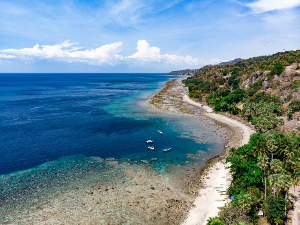  Praia em Adara na ilha de Ataúro em Timor Leste, mar muito claro, areai branca, muitos coqueiros, quatro barcos no mar e algumas montanhas ao redor
