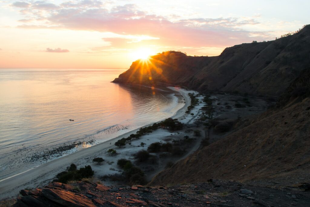 Díli, a capital do Timor Leste, uma região com montanhas, praia e vegetação se misturando na areia da praia, com o sol se pondo atrás de uma das montanhas, para representar o seguro viagem para Timor Leste