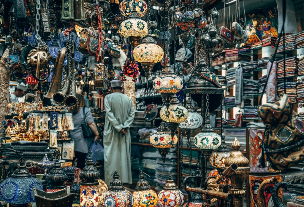 Mercados de rua em Mascate, na capital de Omã, diversos lustres e enfeites para casa em tons de amarelo, verde, com desenhos árabes, muitos tapetes, e duas pessoas observando o local, para representar o seguro viagem para Omã