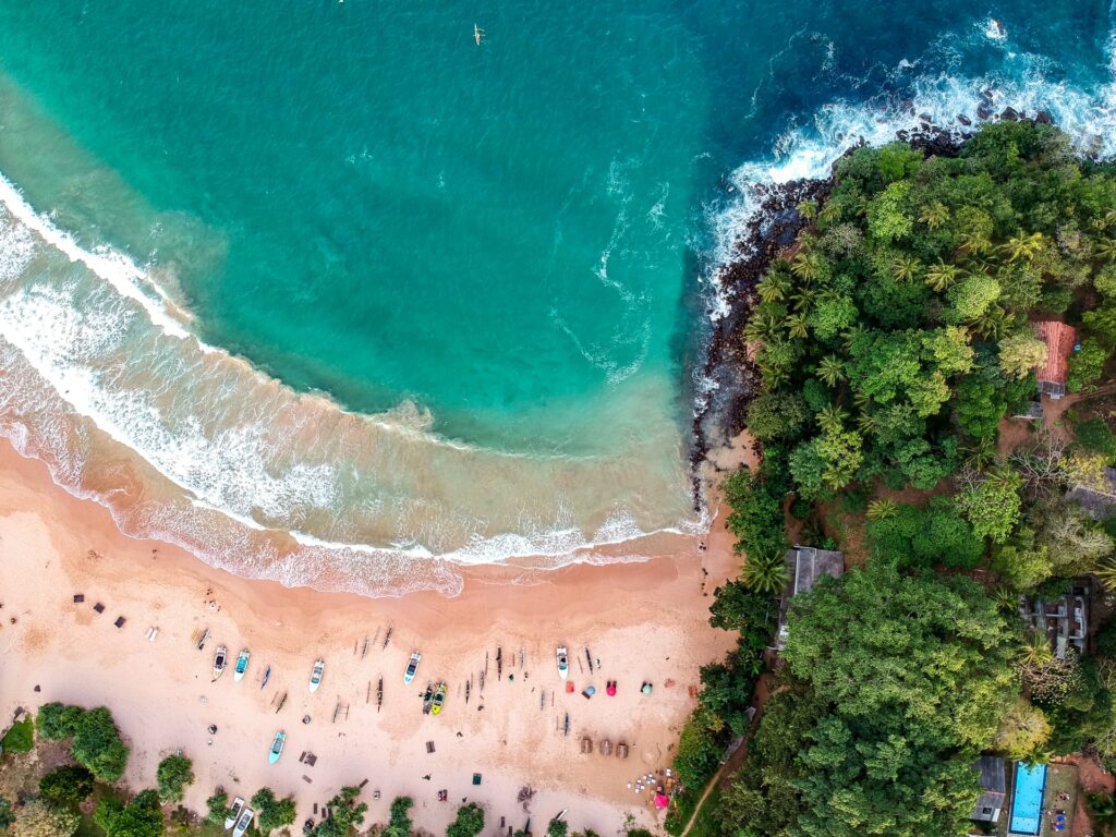 Vista de cima de uma praia com pessoas na areia, muitas árvores e vegetação ao redor e o mar em tom de verde claro, para representar o seguro viagem para o Sri Lanka