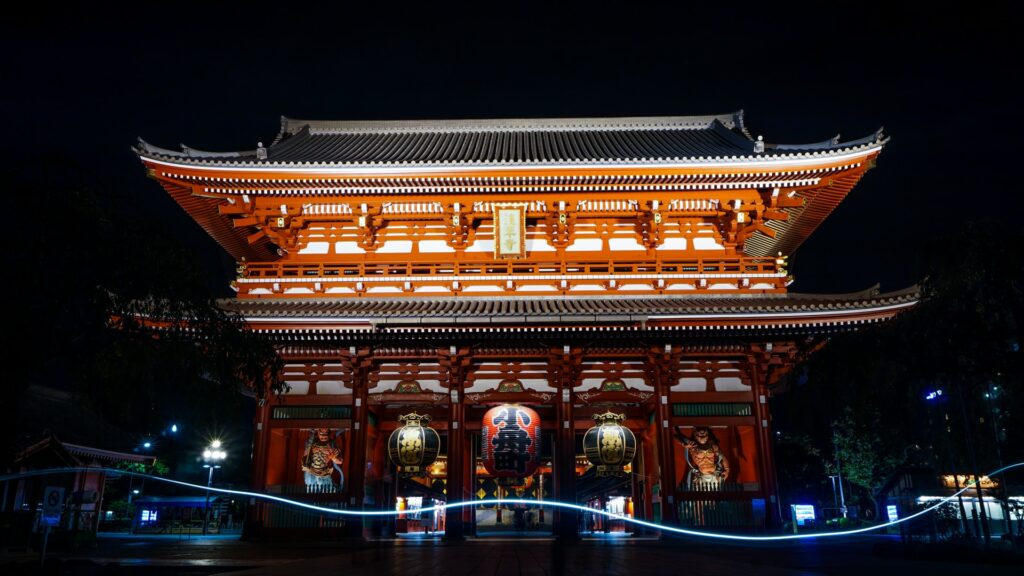 Templo de Asakusa, inteiro aceso de noite, um templo com arquitetura oriental, em tons de vermelho e verde, com figuras budistas logo na entrada do local, para representar o seguro viagem para Tóquio