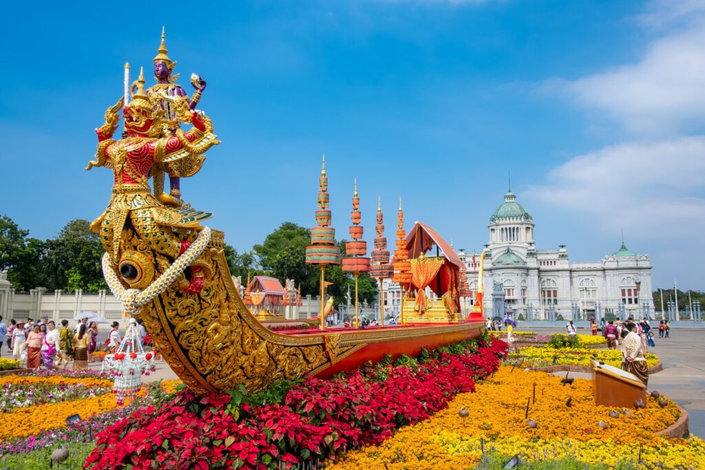 Um enorme barca real banhada em ouro, com símbolos da cultura tailandesa, cercado por flores coloridas em um pátio com pessoas circulando ao redor, para representar o seguro viagem para Tailândia