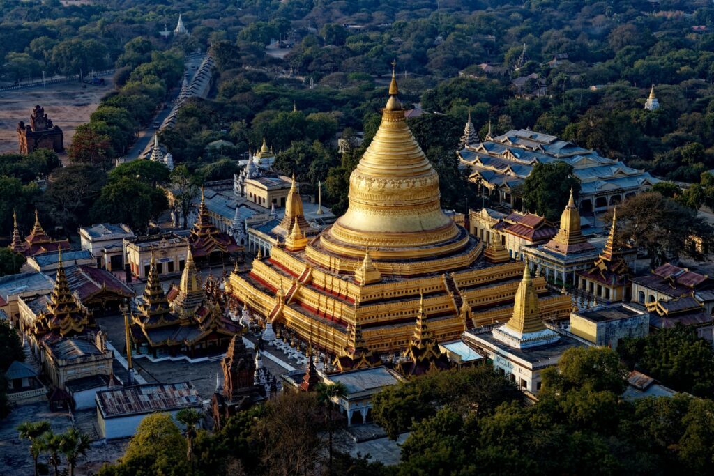 Pagode Shwedagon em Mianmar, um templo budista bem amplo em cor de ouro, com uma cúpula central enorme, cercado por outras pequenas torres, muita vegetação e montanhas ao redor, para representar o seguro viagem para Mianmar