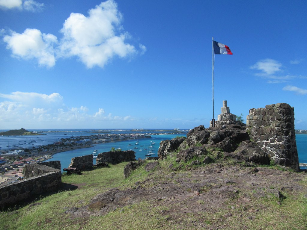 Forte Francês em St Martin, uma construção antiga com rochas e a bandeira da França estiada, após o forte é possível ver o mar e algumas embarcações
