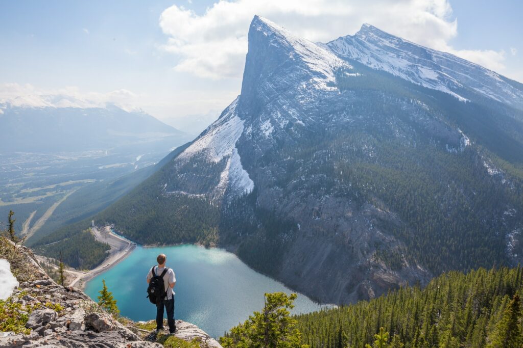 Homem com mochila no alto de uma montanha, olhando para paisagem com um lago de águas claras e pinheiros, ilustrando o post My Travel Assist
