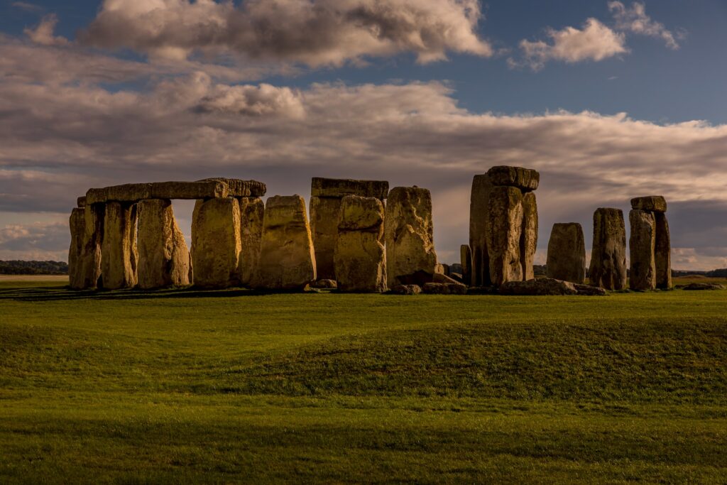 vista do Stonehenge, um monumento com pedras retangulares colocadas retas e algumas em cima das outras, em um gramado verde baixo e céu azul com nuvens, sendo um marco da Inglaterra