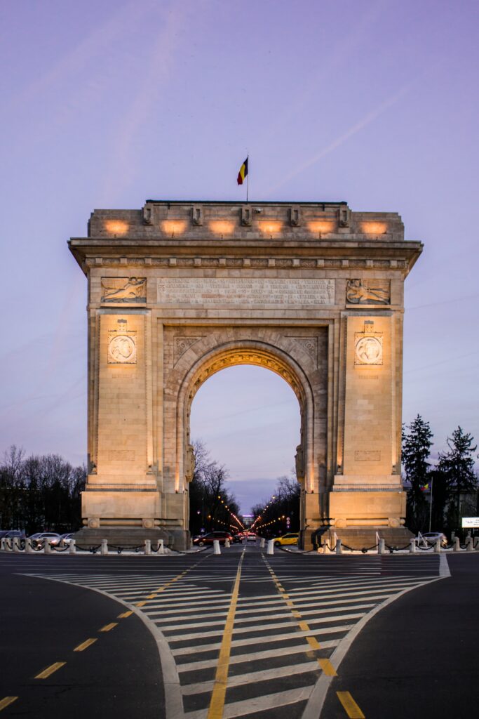 vista do Arco do Triunfo ao entardecer, com luzes do arco e a via abaixo, acima, a bandeira da Romênia, que pode ser visitado com um seguro viagem para Bucareste