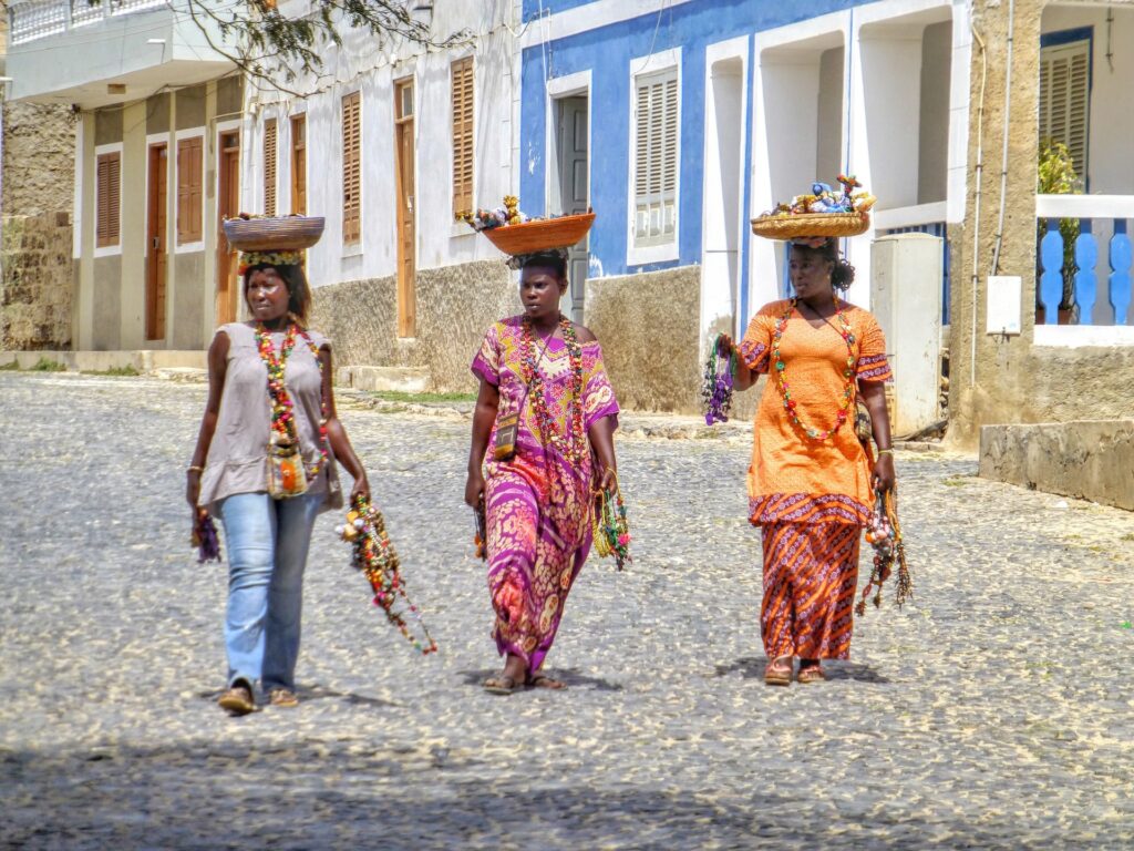 Três mulheres andando em uma rua de pedra, com casinhas coloridas, elas são vestidas com roupas coloridas também e carregando cestas  sob as cabeças, para representar seguro viagem Cabo Verde