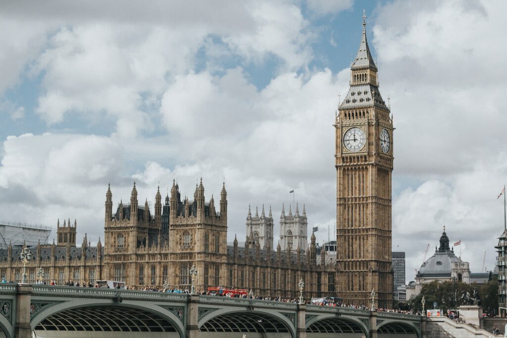 Torre do relógio Big Ben para ilustrar o post sobre chip internacional para o Reino Unido. Vária pessoas passeiam pelaa ponto ao lado, e há um palácio ao lado. A foto foi tirada durante o dia e o céu está azul com nuvens. - Foto: Marcin Nowak via Unsplash