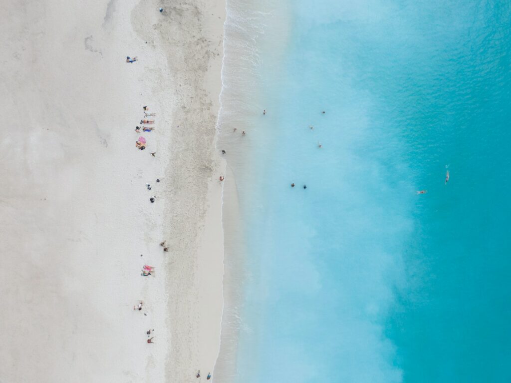 Vista de cima de uma praia com mar muito azul e bem claro, areia branca e com pessoas no local
