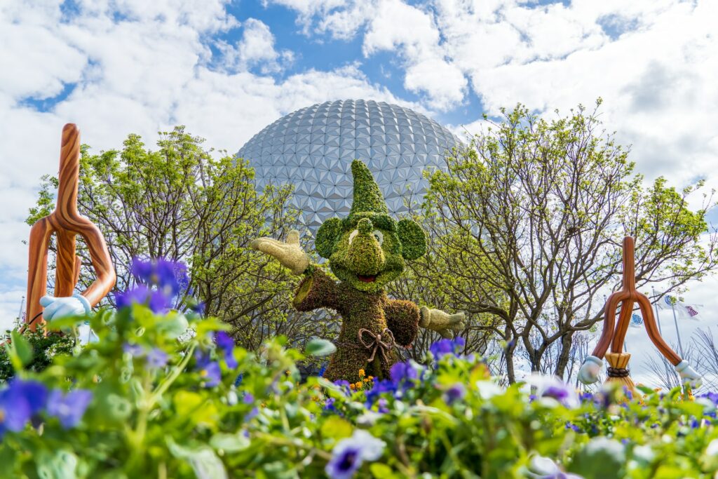 Epcot, um dos parques que pode ser visitado com o chip internacional para a Disney, com o Mickey podado na grama com chapéu de mágico, flores roxas e árvores baixas ao redor, ao fundo, a famosa bola