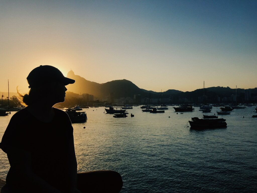 Uma mulher sentada, em um local no Rio de Janerio, usando um boné observando o mar de perto com diversos barcos e algumas montanhas ao fundo, para representar o seguro viagem nacional