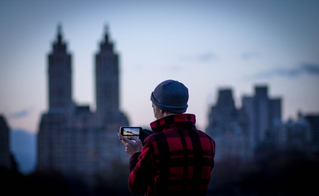 Uma homem com uma jaqueta xadrez vermelha e um toca cinza segurando um celular na horizontal enquanto fotografa a cidade de Nova York