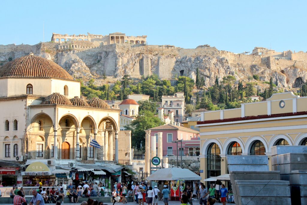 Vista da praça de Monastiraki, em Atenas, durante o dia com várias pessoas andando. Ao fundo, há casas e, mais ao fundo, ruinas históricas. Representa chip internacional para Atenas.
