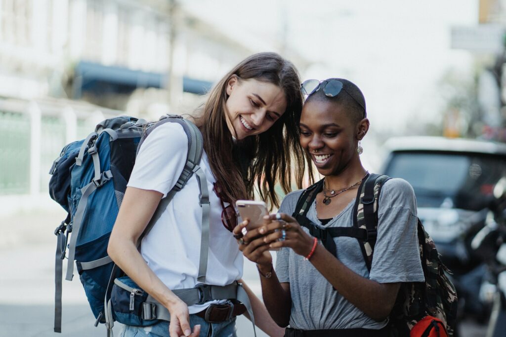 Duas mulheres sorrindo com mochila nas costas e olhando para um celular em que uma delas está segurando. Está de dia e ao fundo, embaçado, tem um carro e uma moto estacionados na rua. Imagem para ilustrar o post chip internacional Toronto