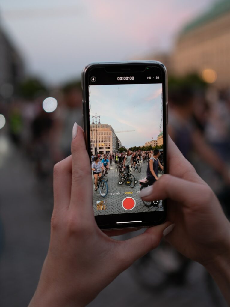Uma mão feminina com unhas brancas segurando um celular na vertical enquanto fotografa uma rua com pessoas andando de bicicleta em Berlim