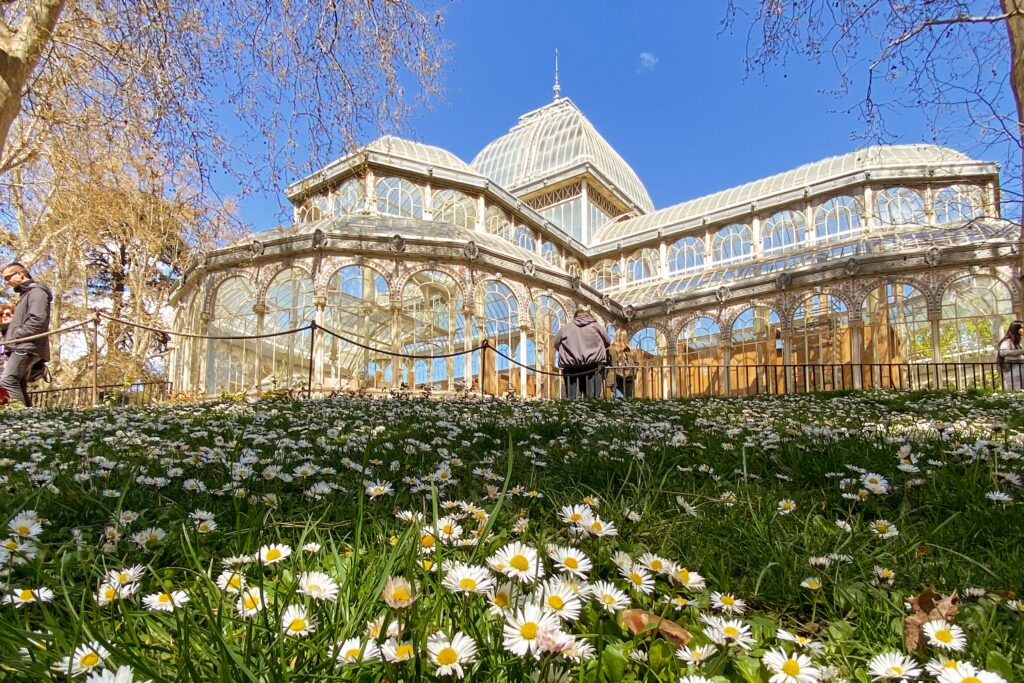 Vista do Palácio de Cristal, Madri, durante o dia, em volta de um jardim com gramado verde, com flores margarida e ao centro um palácio de vidro. Representa chip internacional para Madri.