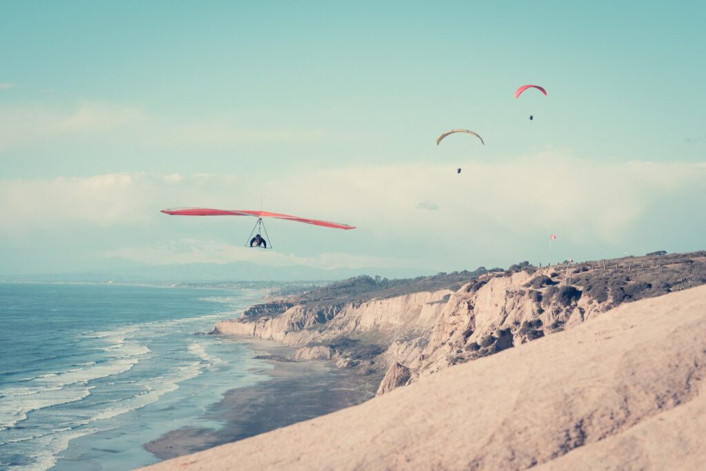 Três pessoas voando de parapente no céu acima de uma praia para ilustrar o post sobre chip internacional para San Diego. Há colinas de um lado, e o mar está do outro. - Foto: Lance Anderson via Unsplash