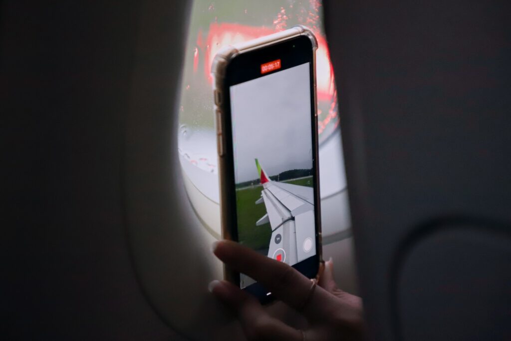 Uma mão feminina segurando um celular na vertical próximo de uma janela do avião enquanto fotograva a asa do avião