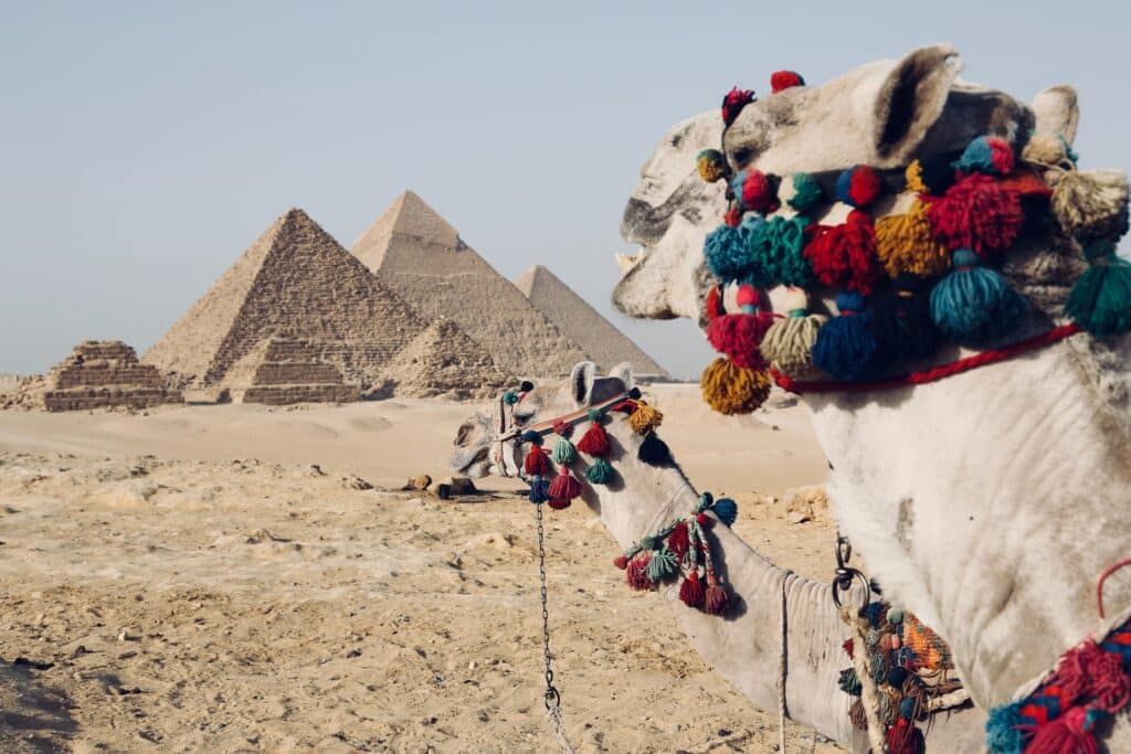 vista das três Pirâmides de Gizé no meio do deserto, com algumas menores em formato de escada abaixo e, na frente, dois camelos com ornamentos coloridos em bolinhas vermelhas, amarelas e azuis, que pode ser visitado com o auxílio de um seguro viagem Cairo