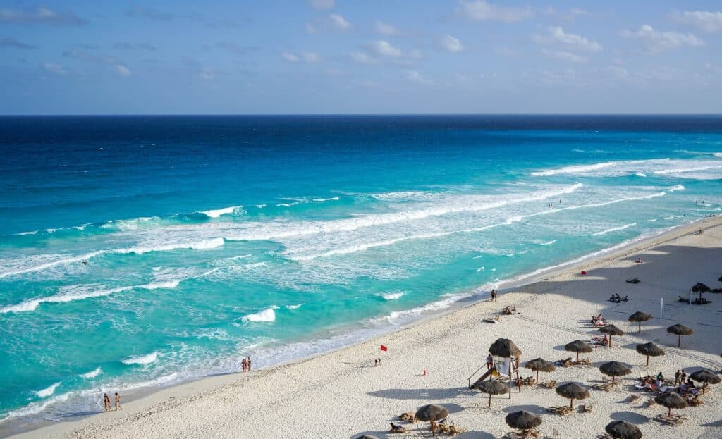 Foto aérea de praia para ilustrar o post sobre chip internacional para Cancun. Há alguns guarda-sóis na areia branca, e pessoas caminham na faixa de areia perto do mar. - Foto: Michelle_Maria via Pixabay