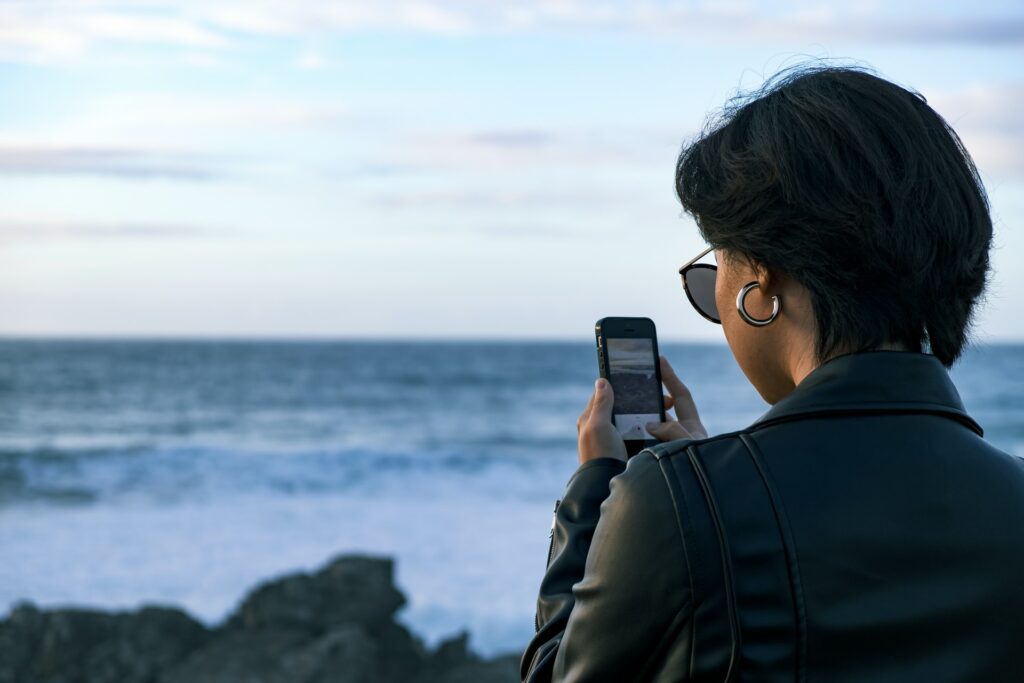 Uma mulher de costas de cabelo curto preto, usando uma jaqueta de couro preta enquanto segura um celular na vertical e fotografa uma praia que está na sua frente