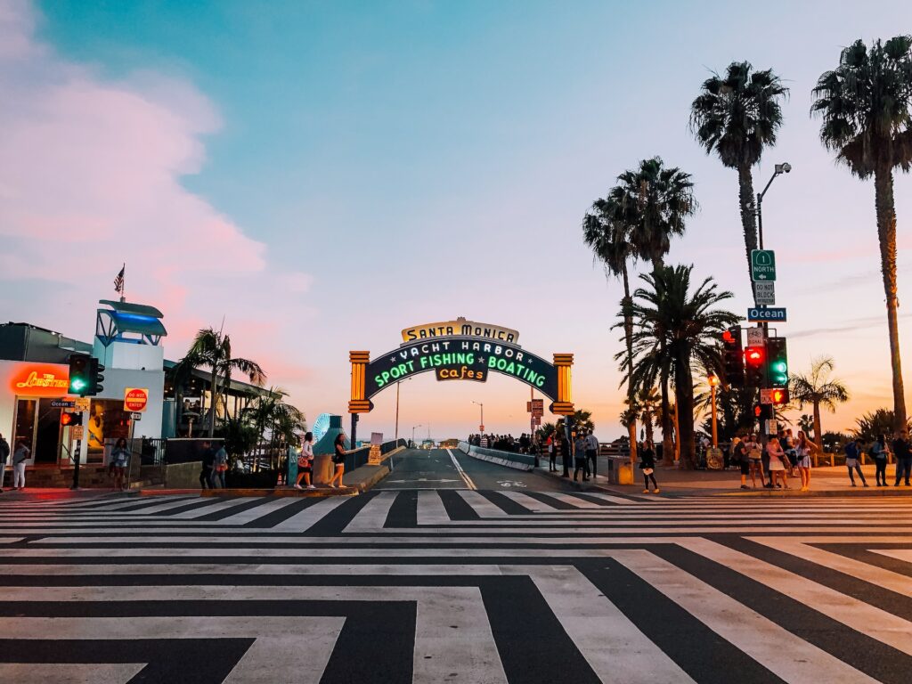 Entrada para o pier de Santa Monica para ilustrar o post sobre chip internacional para Los Angeles. Há uma placa indicando o lugar, e várias pessoas caminham ao redor. Faixas de pedestre ocupam a rua à frente. A foto foi tirada de dia. - Foto: Gerson Repreza via Unsplash