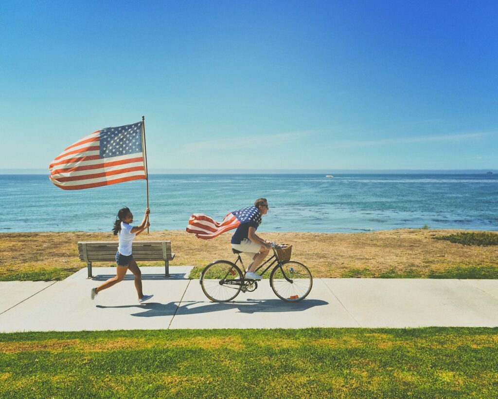 Homem andando de bicicleta com uma bandeira dos EUA nas costas, sorrindo, enquanto uma menina corre atrás segurando outra bandeira dos EUA com as duas mãos. Eles estão em uma ciclovia, com gramado baixo nos lados e ao fundo uma praia de San Diego, num dia ensolarado